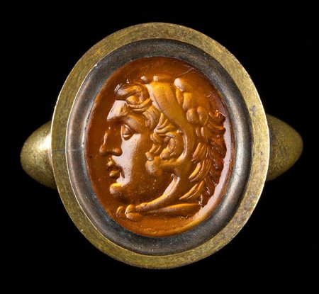 AN ORANGE GLASS IMPRESSION SET IN A GOLD RING. HEAD OF ALEXANDER THE GREAT. 