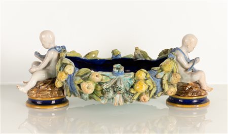 Centrotavola di gusto robbiano neoquattrocentesco con ai lati due putti in maiolica bianca modellati a tutt tondo che trattengono una ghirlanda di frutta e fiori formante le pareti dell'oggetto dipinto all'interno in blu di Prussia   